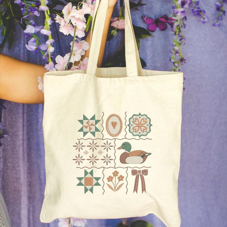 May include: Cream-colored tote bag with a patchwork design featuring geometric shapes, a heart, flowers, and a duck. The bag has two handles and is held up against a purple background with flowers.