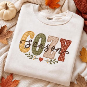 May include: A cream-colored crewneck sweatshirt featuring the word COZY in large, multi-colored plaid block letters and the word season in dark brown cursive script. Below the text is an embroidered design of green leaves, yellow berries, and a small red heart.