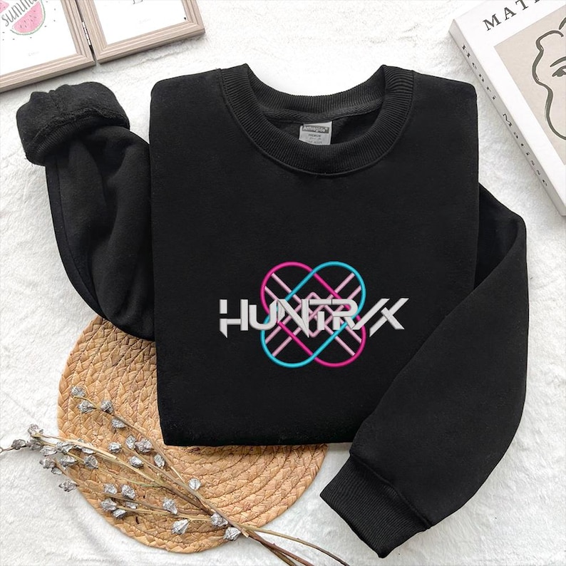 Puede incluir: Sudadera negra con la palabra "HUNTRXX" en blanco, con un dise&ntilde;o de s&iacute;mbolo de infinito rosa y azul. La sudadera est&aacute; sobre una esterilla tejida con una ramita decorativa de flores secas.