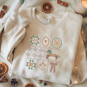 May include: Cream-colored sweatshirt with a patchwork design featuring geometric shapes, a heart, a duck, flowers, and a bow. The design incorporates shades of green, orange, and brown. The sweatshirt is made of a soft material.