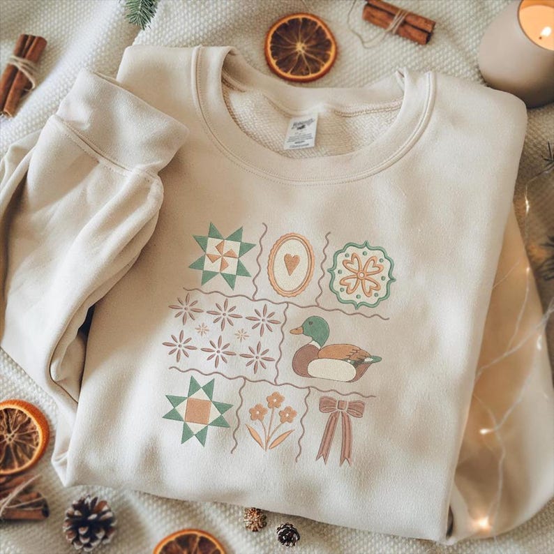 May include: Cream-colored sweatshirt with a patchwork design featuring geometric shapes, a heart, a duck, flowers, and a bow. The design incorporates shades of green, orange, and brown. The sweatshirt is made of a soft material.