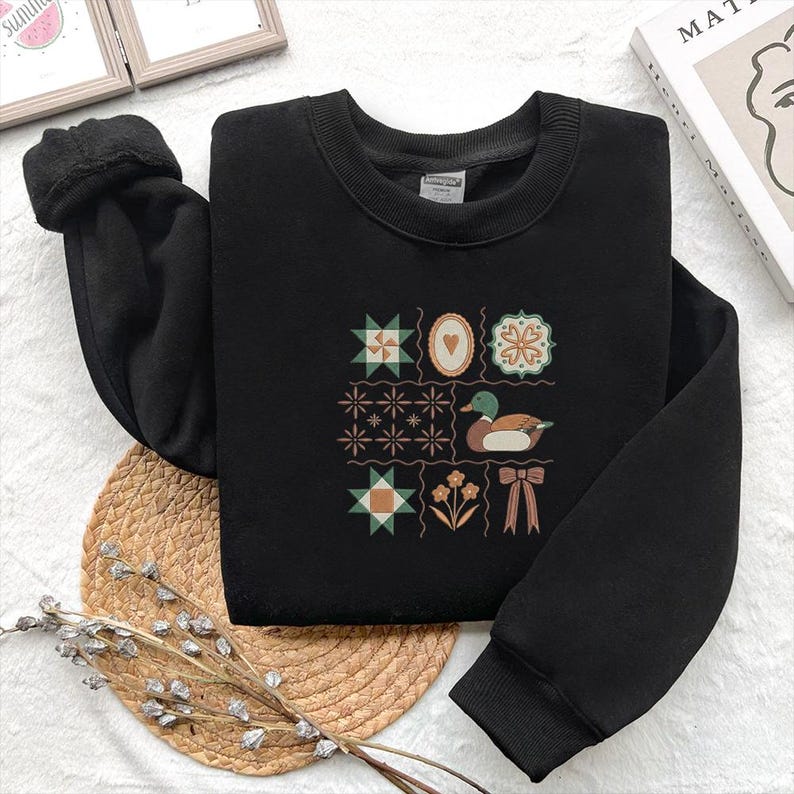 May include: Black sweatshirt with a crew neck and long sleeves. The front features a patchwork design with a duck, flowers, and a bow. The design includes geometric shapes in green, brown, and orange.