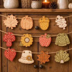 May include: A decorative garland featuring three rows of fabric cutouts in autumn-themed shapes including leaves, pumpkins, acorns, mushrooms, sunflowers, apples, sweaters, hats, and turkeys. Each piece is stitched with various patterns and colors.