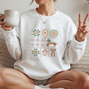 May include: White sweatshirt featuring a patchwork design with a duck, heart, flowers, and geometric shapes. The design includes shades of green, orange, and brown. The sweatshirt has a crew neck and long sleeves.