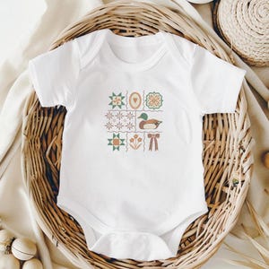 May include: White baby onesie with a patchwork design featuring a duck, flowers, and geometric shapes in shades of green, brown, and orange. The onesie is short-sleeved and has a round neckline. The garment is displayed in a woven basket.