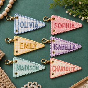May include: Six triangular fabric pennant keychains with gold-tone lobster clasps and metal grommets. Each features embroidered names: Olivia on blue gingham, Sophia on pink polka dots, Emily on yellow gingham, Isabella on purple gingham, Madison on light blue, and Charlotte on a star print.