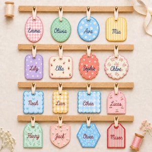 May include: Sixteen fabric tags in various shapes and patterns, each featuring a name embroidered in dark thread. The tags are suspended by cream-colored ribbons from four horizontal wooden bars. Patterns include gingham, polka dots, floral, and stripes.