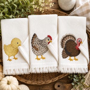 May include: Three off-white waffle-weave kitchen towels with fringe edges. The left towel features an embroidered yellow chick, the center towel shows a speckled brown and white hen, and the right towel displays a brown turkey with red accents.