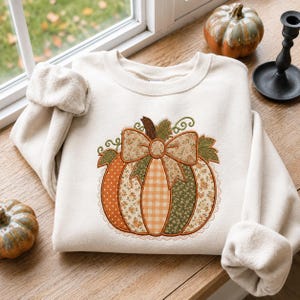 May include: A cream-colored crewneck sweatshirt featuring a central embroidered pumpkin graphic. The pumpkin is composed of various fabric-like patterns in orange, green, and beige, topped with a large bow and surrounded by green leaves.