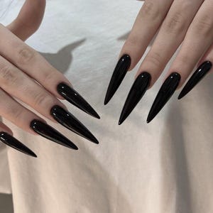 以下が含まれることがあります： 光沢のあるソリッドブラック仕上げの、長く尖ったスティレット型の爪。爪は鋭く細長い先端に整えられており、暗く不透明な色の上に光を反射する滑らかな表面が特徴です。