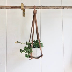 Puede incluir: Una maceta de cerámica blanca con una planta suculenta colgando de un macetero de macramé de cuero marrón.