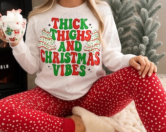 Puede incluir: Una sudadera blanca de manga larga con el texto en rojo y verde "THICK THIGHS AND CHRISTMAS VIBES" y dos árboles de Navidad ilustrados, combinada con pantalones de pijama rojos con lunares blancos y zapatillas de estar por casa beige.