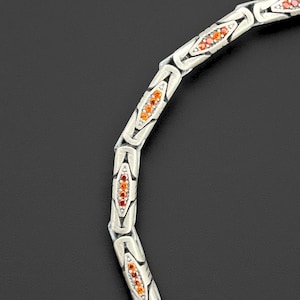 May include: A silver-toned necklace with a repeating pattern of elongated, gem-filled links. Each link is set with small, orange-red gemstones. The necklace is displayed against a dark background, highlighting its intricate design and color.