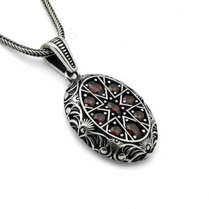 Puede incluir: Collar con colgante ovalado de plata con un diseño floral detallado y un patrón de estrella de piedras preciosas rojas. El colgante cuelga de una cadena de plata, creando un aspecto clásico y elegante.