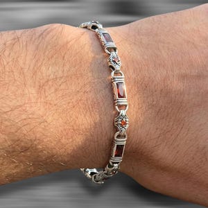 Puede incluir: Una pulsera de eslabones de plata con piedras preciosas rectangulares de color rojo intenso que se alternan con piedras circulares más pequeñas de color naranja. Los eslabones metálicos tienen grabados detallados.