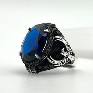 May include: A silver ring featuring a large, oval-shaped blue gemstone. The gemstone is encircled by smaller black stones. The ring has detailed patterns on the sides, with an ornate design. The ring is displayed against a white backdrop.