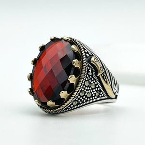 Könnte beinhalten: Ein silberner Ring mit einem großen, facettierten, ovalförmigen roten Edelstein. Der Ring hat goldfarbene Akzente um den Edelstein und am Band, mit aufwendigen Details. Das Design ist verziert und hat eine kronenartige Fassung für den Stein.