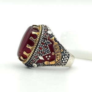May include: A silver ring with a large, oval, deep red stone. The ring features gold accents around the stone and on the sides, with intricate detailing and a crown-like design. The ring is set against a white background.