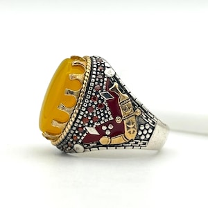 May include: A silver ring with a large, oval, yellow gemstone. The ring features intricate detailing on the band, including gold accents, red enamel, and small gemstones. The design incorporates geometric and symbolic elements.