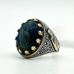 May include: A silver ring with a large, oval, blue gemstone. The ring has gold-colored accents around the stone and on the sides, with a detailed pattern. The gemstone has a striped appearance.