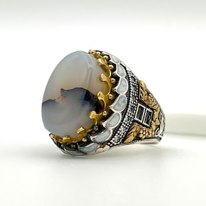 May include: A silver ring featuring a large, oval, pale grey stone with dark grey and brown markings. The ring has gold-coloured detailing around the stone and on the band, with small white and black embellishments. The ring is set against a white backdrop.