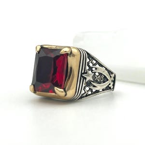 Könnte beinhalten: Ein silberner Ring mit einem großen, rechteckigen, tiefroten Edelstein. Die Fassung ist goldfarben um den Stein, und das Band hat ein dekoratives Design mit einem Stern- und Halbmondmotiv.