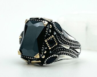 May include: A silver ring with a large, rectangular black gemstone. The ring has intricate detailing around the stone and on the band, with gold-colored accents. The design is ornate and eye-catching.