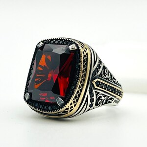 Könnte beinhalten: Ein silberner Ring mit einem großen, rechteckigen, tiefroten Edelstein. Der Ring hat aufwendige Details, darunter schwarze Akzente um den Edelstein und goldfarbenes Metall an den Seiten. Das Band weist verzierte Muster auf.
