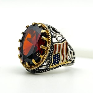 Op de afbeelding: Zilveren ring met een grote, ovale, rode edelsteen gezet in een goudkleurige bezel. De ring heeft een Amerikaans vlagontwerp aan de zijkant en ingewikkelde details. Een opvallend sieraad.