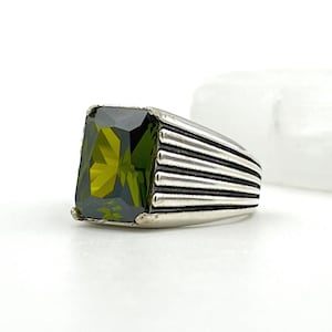 May include: A silver ring with a large, rectangular, olive-green gemstone. The band has a ribbed design. The gemstone is faceted, reflecting light. The ring is set against a white background.