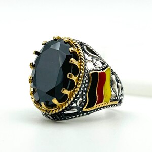 May include: A silver ring with a large, oval-cut black gemstone. The ring features gold-colored accents around the stone and a decorative band with the German flag. The ring is set against a white background.