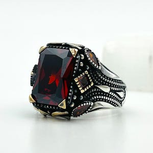 May include: A silver ring with a large, rectangular, deep red gemstone as the centerpiece. The ring features gold accents and smaller, reddish-brown stones. The band has a detailed, textured design.
