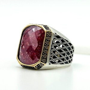 May include: A silver ring with a large, faceted, burgundy-colored stone. The stone is set in a gold-colored bezel. The ring band features a black, patterned design on the sides.