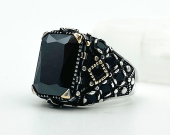 May include: A silver ring with a large, rectangular black gemstone. The ring features intricate detailing with small black and gold accents around the stone and on the band. The ring is set against a white background.