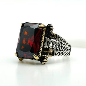 Könnte beinhalten: Ein silberner Ring mit einem großen, rechteckigen, facettierten, dunkelroten Edelstein. Der Ring hat goldfarbene Akzente um den Edelstein und an den Seiten. Das Band hat ein detailliertes, strukturiertes Design.