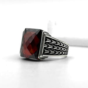 May include: A silver ring with a large, faceted, rectangular, deep red gemstone. The band features a repeating arrow design. The ring is set against a white background, highlighting the jewelry's details and craftsmanship.
