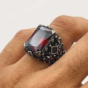 May include: A silver ring featuring a large, rectangular, faceted deep red gemstone set in the centre. The band is crafted with intricate, dark, textured metalwork and smaller red stone accents along the sides of the setting.