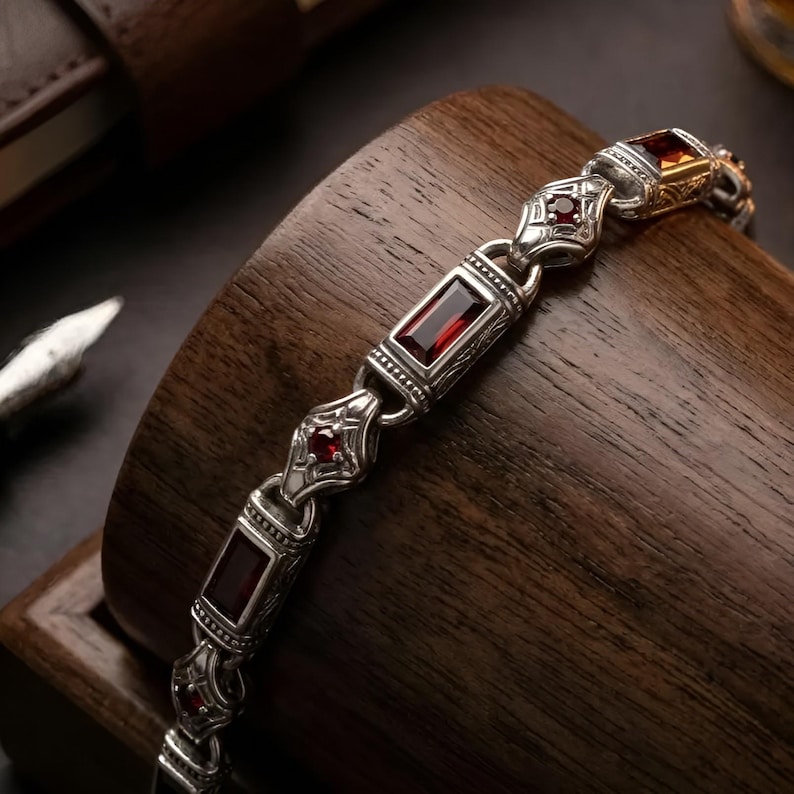 Peut inclure: Un bracelet en m&eacute;tal argent&eacute; compos&eacute; de maillons altern&eacute;s rectangulaires et en forme de losange. Les maillons rectangulaires sont sertis de pierres rouge fonc&eacute; taille baguette, tandis que les losanges contiennent de petites pierres rondes.