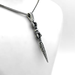 May include: A silver-colored pendant necklace featuring a detailed, ornate dagger-shaped charm. The charm has intricate patterns and a textured surface, suspended from a delicate chain. The pendant is displayed against a white background.