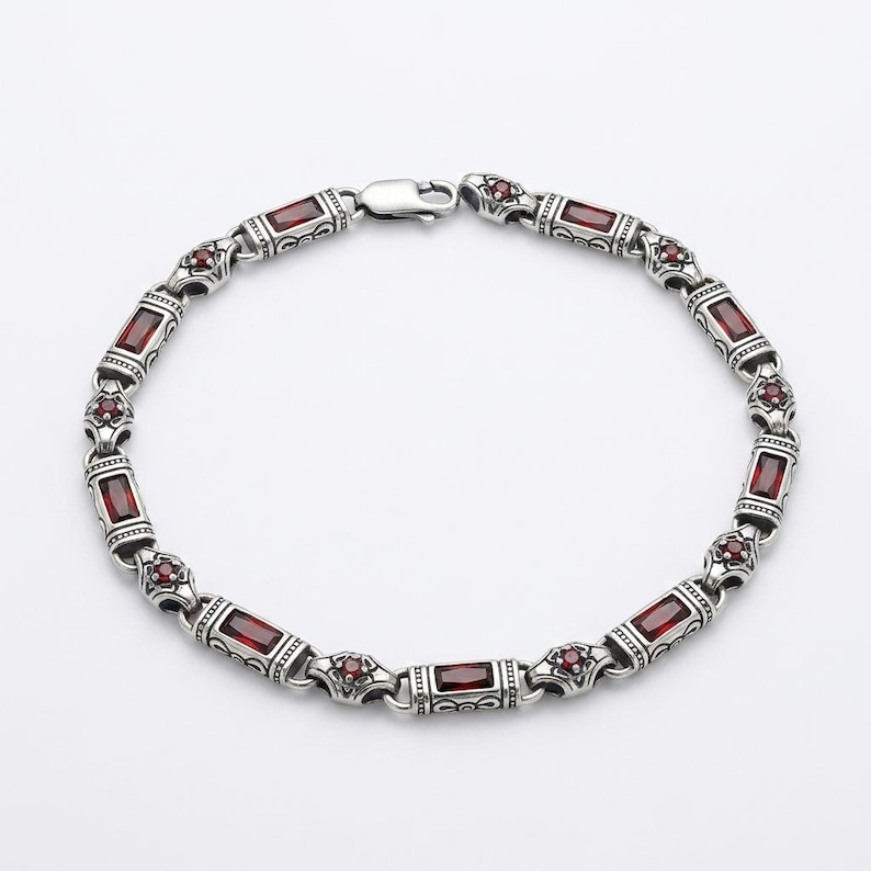 Peut inclure: Un bracelet en m&eacute;tal argent&eacute; compos&eacute; de maillons rectangulaires et circulaires altern&eacute;s. Les maillons rectangulaires sont sertis de pierres rouges fonc&eacute;es taille baguette, tandis que les maillons circulaires contiennent de petites pierres rouges.