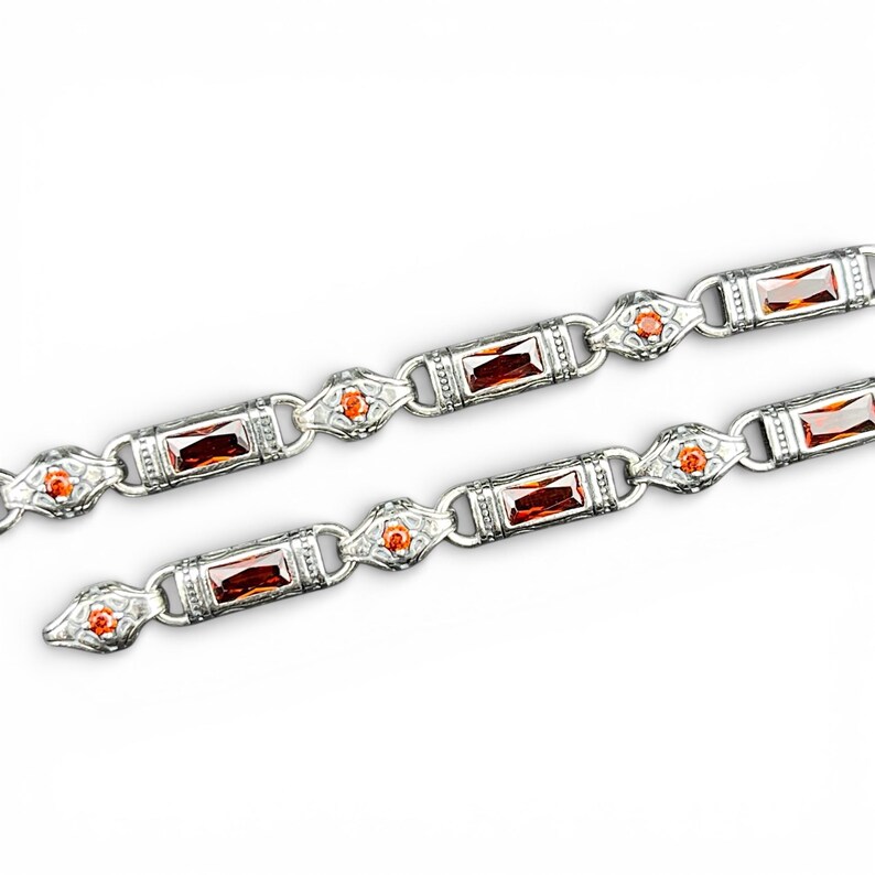 Peut inclure: Un bracelet en cha&icirc;ne argent&eacute;e compos&eacute; de maillons rectangulaires sertis de pierres rouges taille baguette, alternant avec des maillons argent&eacute;s plus petits et orn&eacute;s de petites pierres orange rondes. Le m&eacute;tal pr&eacute;sente des d&eacute;tails textur&eacute;s.