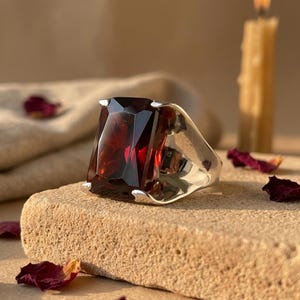 May include: A silver ring featuring a large, rectangular, faceted deep red gemstone. The stone is set in a polished silver band with four prongs securing the corners of the gem. The ring displays a bold, substantial design with a smooth, reflective metal finish.