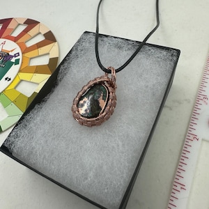 May include: A handcrafted copper wire-wrapped pendant necklace. The pendant features a teardrop-shaped stone with green and brown hues. The necklace is displayed in a black box with white padding, alongside a color wheel and a ruler.