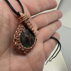 May include: Copper wire-wrapped pendant with a natural stone center. The stone features green and brown hues. The pendant is suspended from a black cord necklace. The wire is intricately woven around the stone, creating a unique, handcrafted design.
