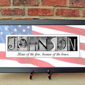 May include: Framed artwork featuring the name "JOHNSON" spelled out in black and white images. The background is an American flag. The text "Home of the free, because of the brave" is below the name. The frame is black.