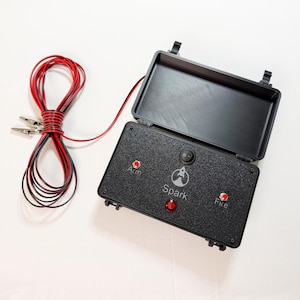 May include: A black rectangular electronic control box with a hinged lid, featuring a rocket icon and the text Spark. The panel includes a central toggle switch, two red push buttons labeled Arm and Fire, and a red indicator light. Red and black wires with metal alligator clips are attached.