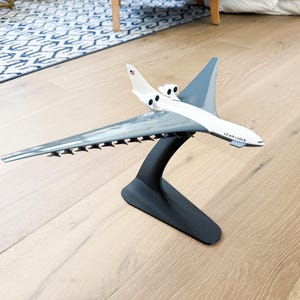 May include: A model airplane, predominantly white and gray, with the text "U.S. AIR FORCE" on the nose, is displayed on a black stand. The wings are gray, and the tail features the American flag. The model sits on a light wood floor.