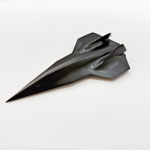May include: A sleek, black sculpture of a futuristic aircraft. The design features a long, pointed nose and a streamlined body with a fin on top. The object is set against a plain white background, highlighting its form and shape.