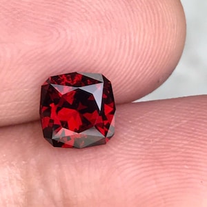 May include: A faceted, cushion-cut gemstone with a deep red hue. The gem has a complex internal structure, reflecting light to create a brilliant sparkle. The stone is held between fingers, showcasing its size and color.