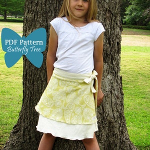 Puede incluir: Una niña pequeña con una camiseta blanca y una falda de envoltura de paisley amarillo y blanco. Está de pie frente a un árbol y tiene una flor blanca en el pelo. El texto "PDF Pattern Butterfly Tree" está en forma de mariposa azul.